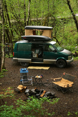 Van life adventure! This setup, like an RV rental, features a green Astro van with a rooftop tent, campfire, and chairs in a lush forest.