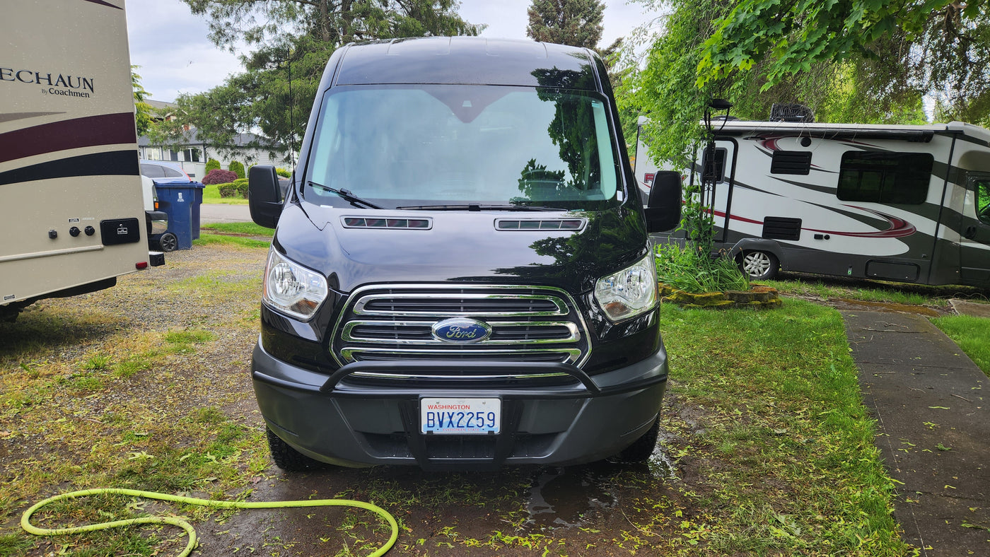 Front view of a black Ford Transit RV rental van (WA plate) parked on grass between two large RVs in a residential setting. — photo 4