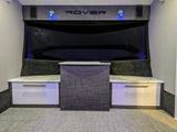 RV rental interior featuring a modern entertainment area with a large dark screen, "ROVER" branding, blue lights, and white/grey cabinets.