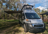 Discover adventure with this pop-top RV rental: a silver camper van with an extended awning at a wooded campsite.