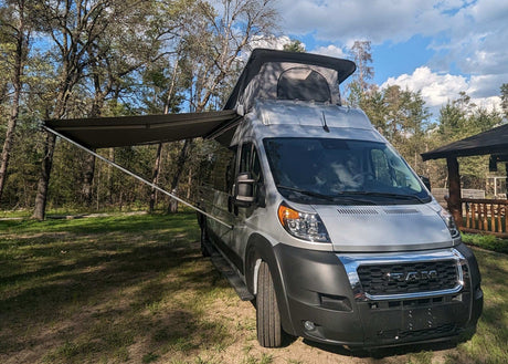 Discover adventure with this pop-top RV rental: a silver camper van with an extended awning at a wooded campsite.