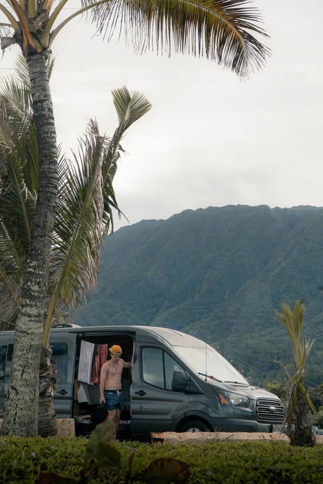 Man in an RV rental van surrounded by tropical nature: palm trees and green mountains.