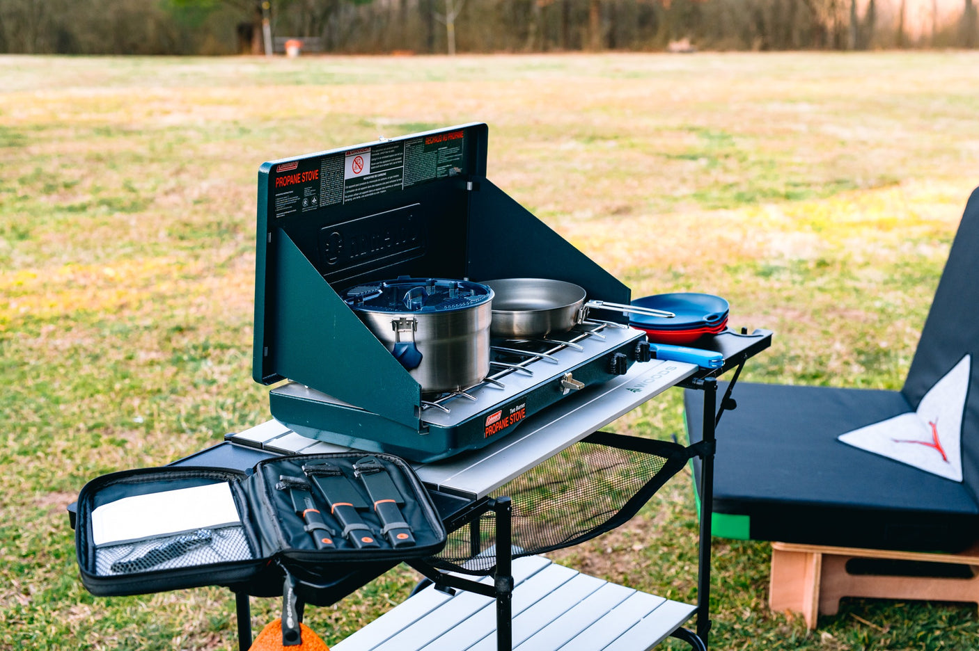 RV rental ready: A Coleman propane stove with pot & pan on a portable camping table, with cooking utensils and plates in an outdoor setting.
