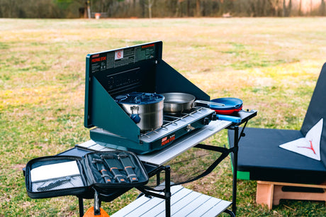 RV rental ready: A Coleman propane stove with pot & pan on a portable camping table, with cooking utensils and plates in an outdoor setting.