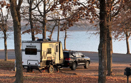 Winnebago Hike RV rental towed by an SUV, parked lakeside under trees with autumn leaves. Ideal for camping.