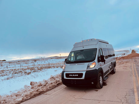 A white Ram rv rental camper van is parked on a snowy road, ideal for winter road trips. Driver waves from inside.