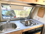 RV rental kitchen: Compact space with sink, stove, kettle, paper towel dispenser, and window view for camping.