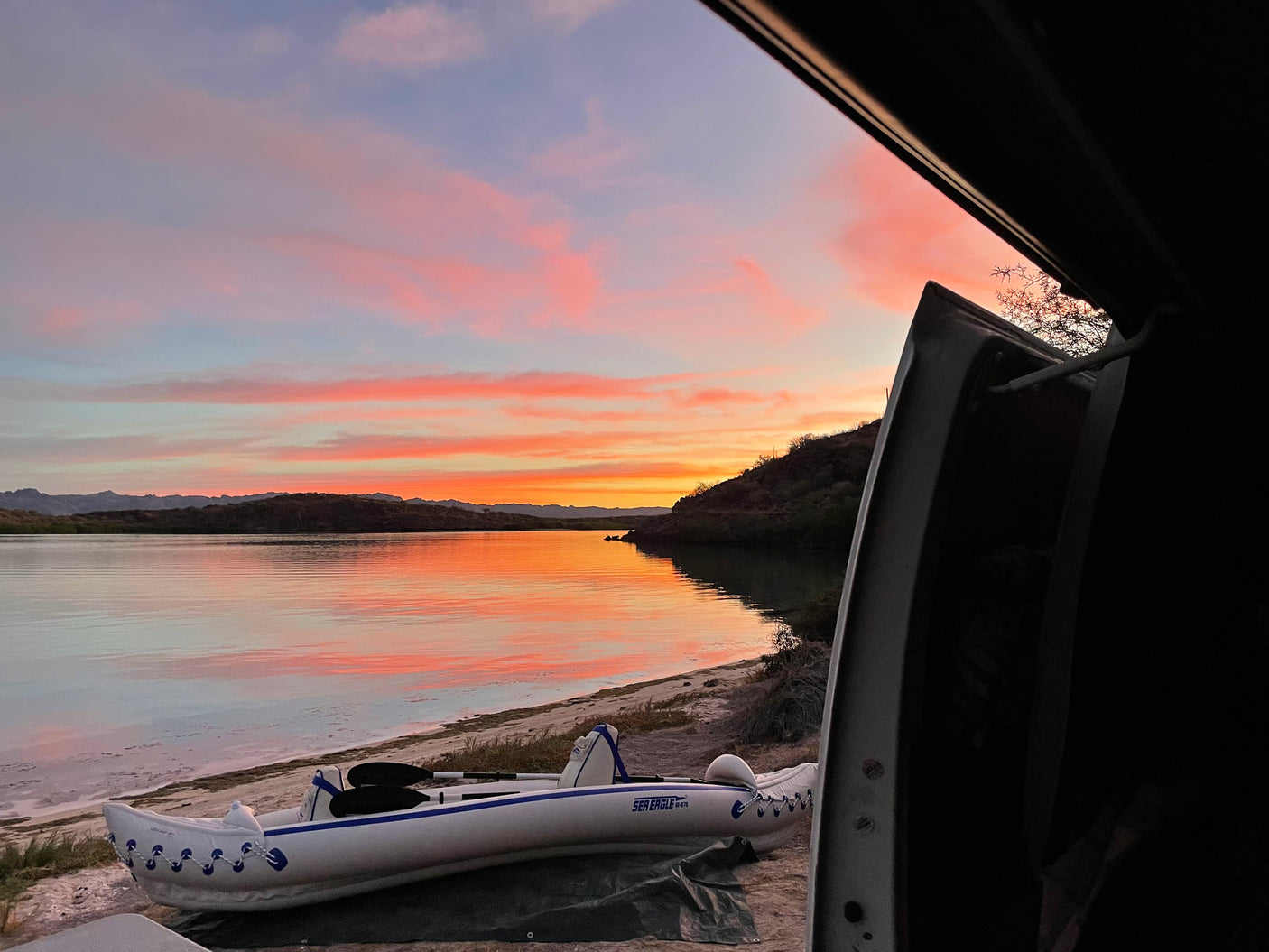 Serene lake sunset from an RV rental doorway, with a beached kayak. Vibrant pink/orange clouds reflect on calm water near mountains.