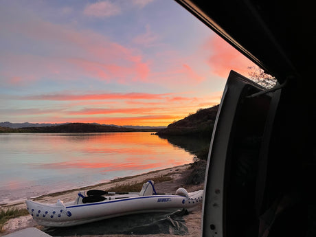 Serene lake sunset from an RV rental doorway, with a beached kayak. Vibrant pink/orange clouds reflect on calm water near mountains.