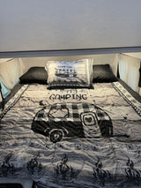 Cozy RV rental bed with "Happy Camping" comforter & pillow, featuring a black & white camper design.