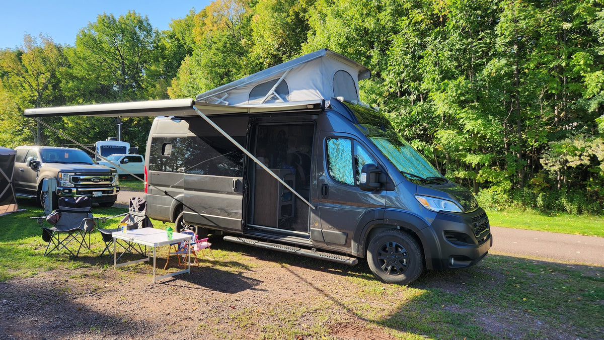 An RV rental, a dark grey camper van with its pop-top and awning out, is set up at a sunny campsite with chairs and a table.