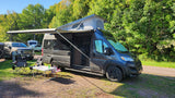 An RV rental, a dark grey camper van with its pop-top and awning out, is set up at a sunny campsite with chairs and a table.