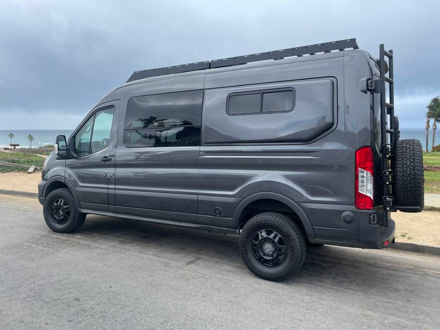 Sleek grey adventure van with roof rack, ladder, and rear tire, parked by the ocean. Perfect RV rental for van life travel.