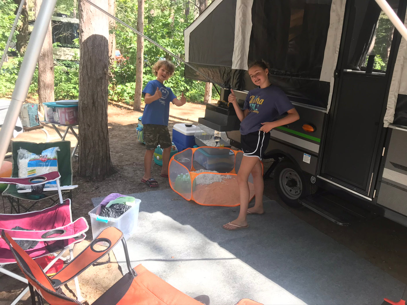 Two happy children give thumbs up next to their RV rental, a pop-up camper, set up at a sunny campsite.