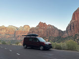 A dark red RV rental van with a roof cargo box is parked on a road, facing majestic red rock mountains.