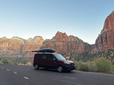 A dark red RV rental van with a roof cargo box is parked on a road, facing majestic red rock mountains.
