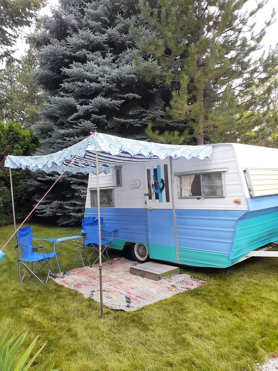 Relaxing RV rental scene: a retro blue & teal camper with an awning, chairs, and rug on green grass.