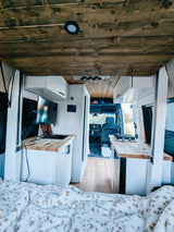 Inside a modern rv rental camper van: rustic wood ceiling, white kitchen with TV, sink, and stove. Cozy blanket in the foreground.