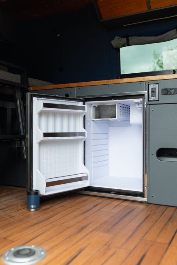 Explore our RV rental kitchen! Bright interior of a compact, open fridge with a small freezer. Wood floor & Yeti cup visible.
