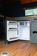 Explore our RV rental kitchen! Bright interior of a compact, open fridge with a small freezer. Wood floor & Yeti cup visible.