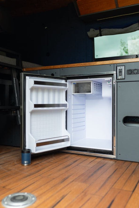 Explore our RV rental kitchen! Bright interior of a compact, open fridge with a small freezer. Wood floor & Yeti cup visible.