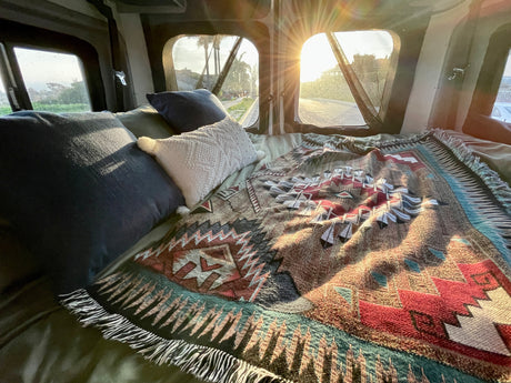 Cozy RV rental bed with pillows & patterned blanket, bright sun shining through windows, offering a relaxing road view outside.