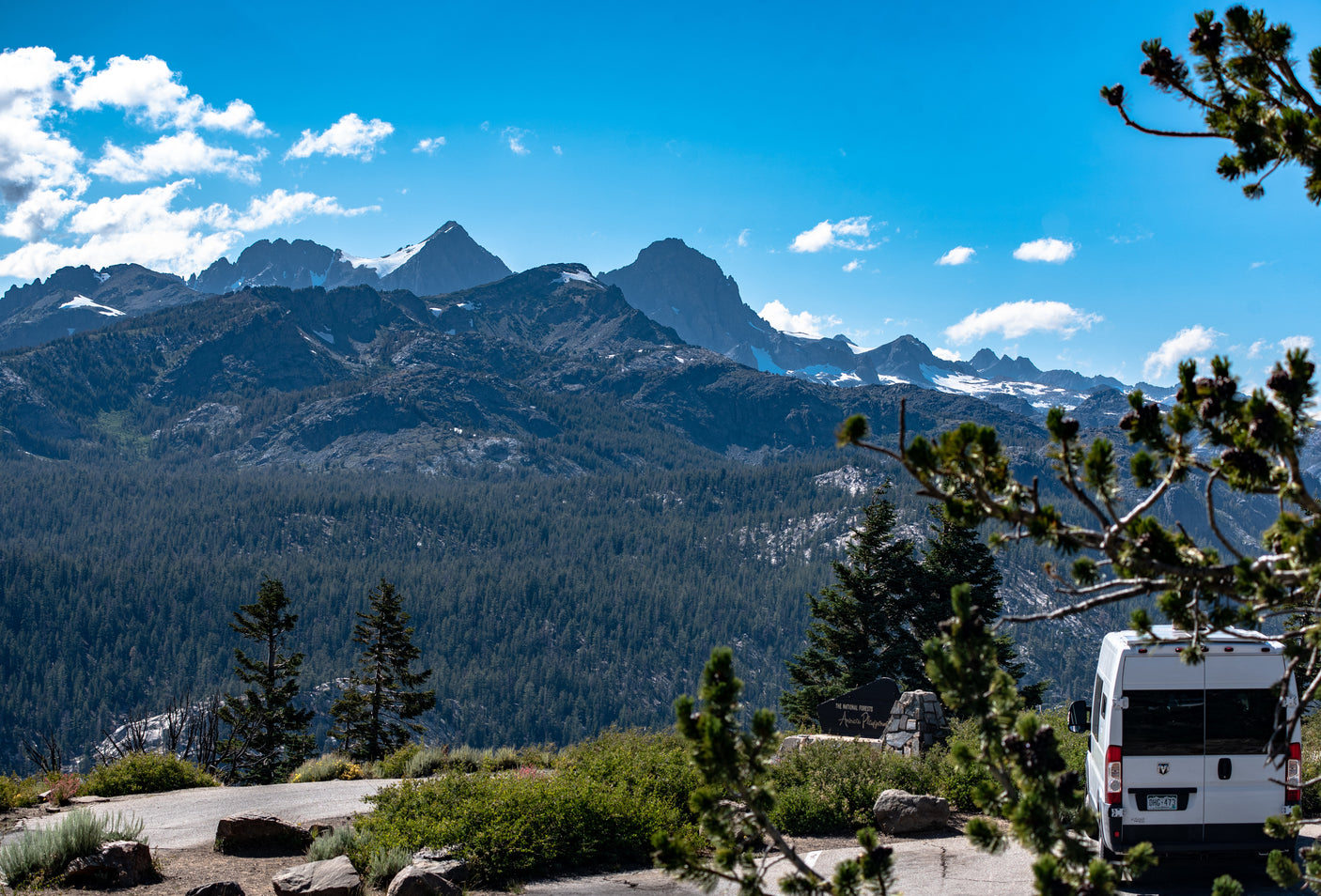 An RV rental van parked at a scenic overlook in a national forest, with majestic snow-capped mountains and a vast forest.