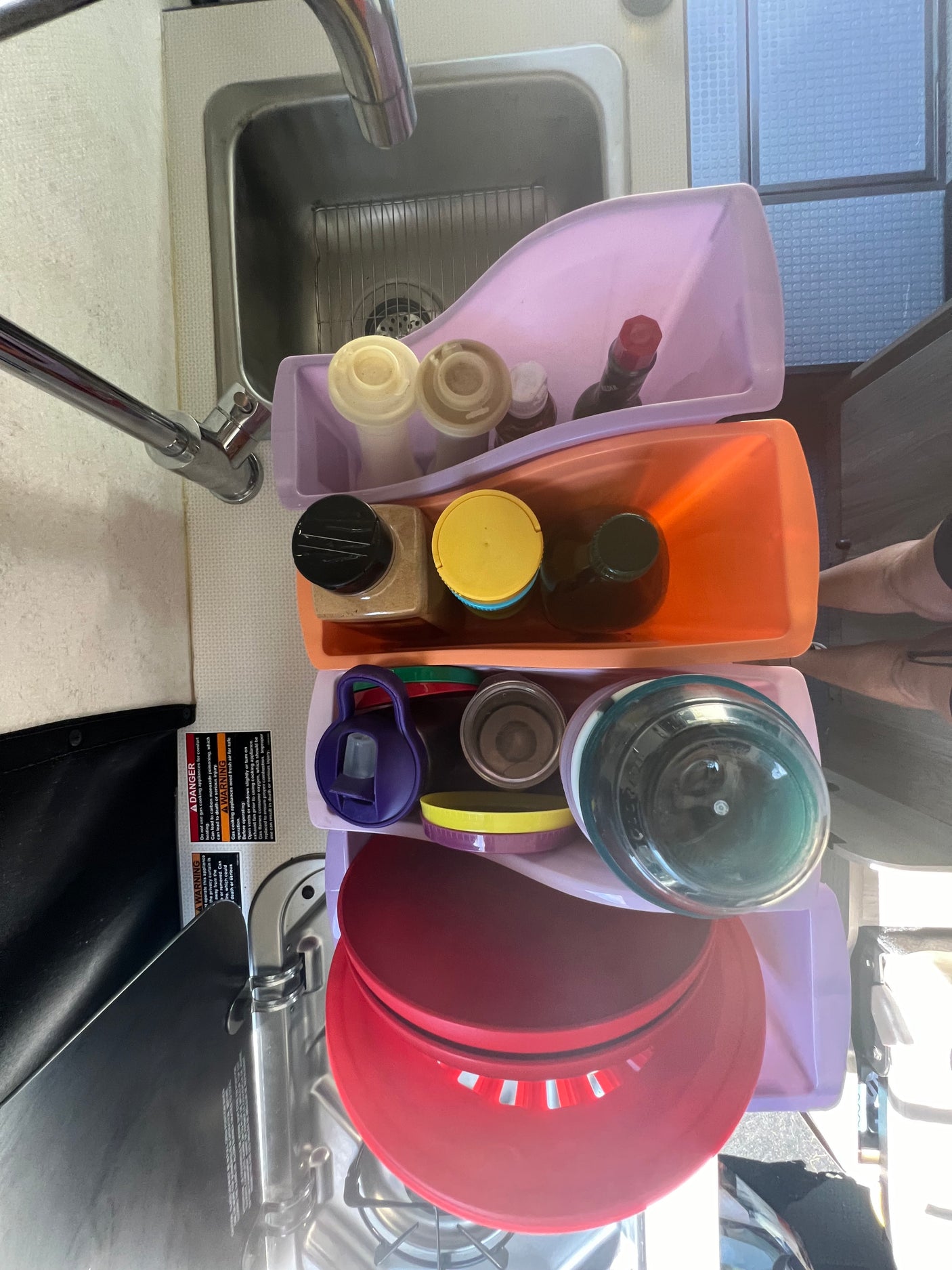 Organized RV rental kitchen sink area with stacked caddies, bottles, and cooking safety warnings visible.