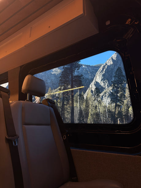Inside this RV rental, rear seats offer a stunning window view of towering grey mountains, evergreen trees, and clear blue sky.