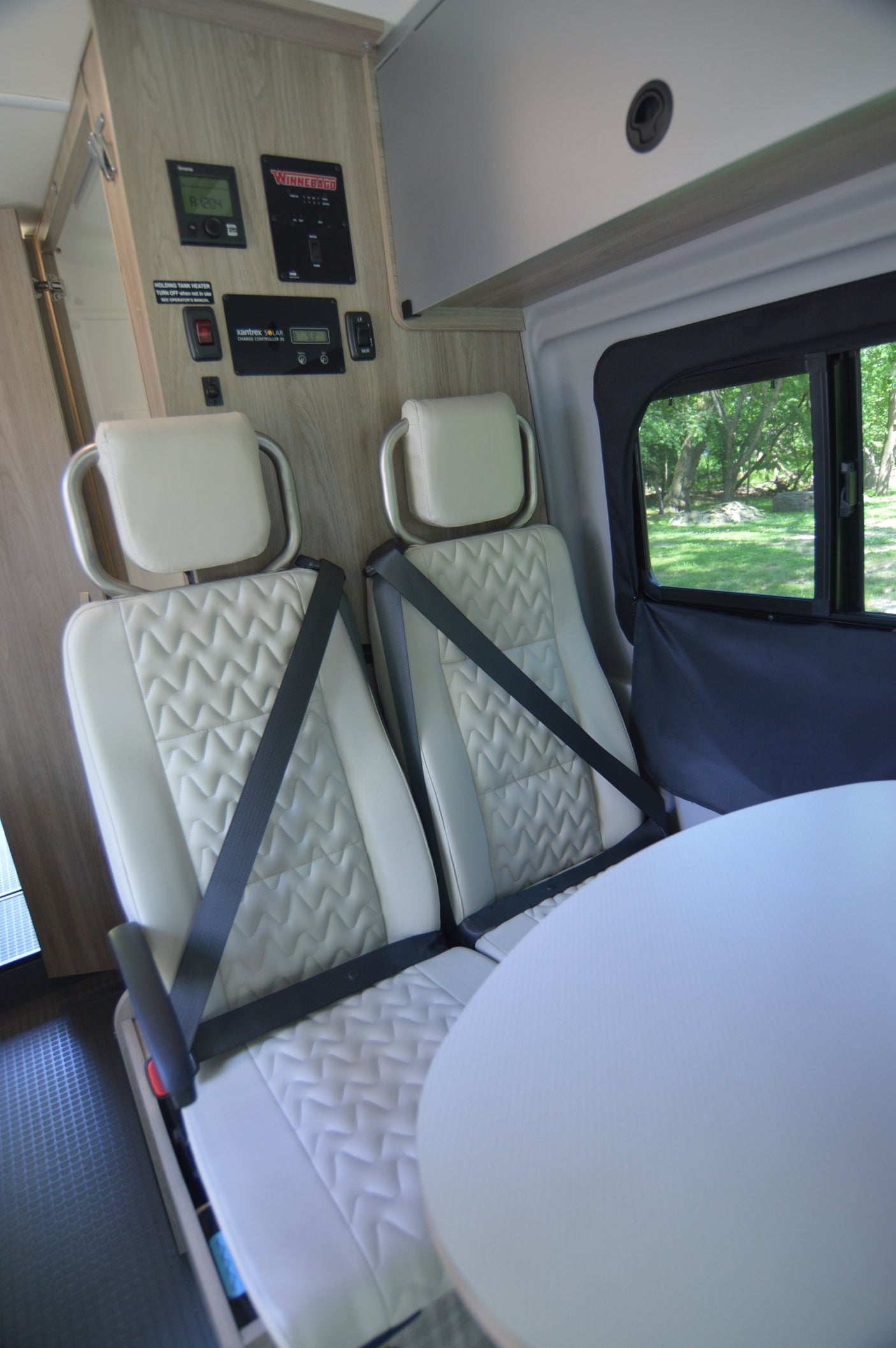RV rental interior view: a dining area with two quilted seats, seatbelts, white table, and control panels on the wall.