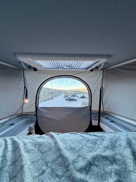 Cozy RV rental interior with a patterned bed, offering a panoramic view of a desert road, parked cars, and distant hills.