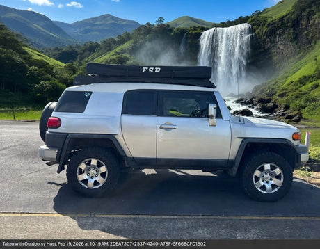 RV rental adventure! Silver Toyota FJ Cruiser with a rooftop tent by a majestic waterfall and green mountains.