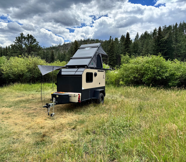 Rugged off-road camper RV rental with rooftop tent, set in a scenic field with forests and cloudy sky. Perfect for adventure.