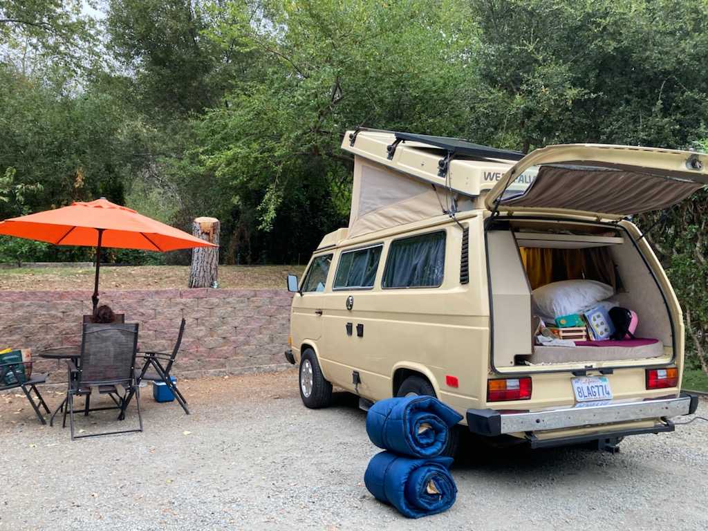  parked on a sunny campsite.|A tan pop-top camper van (RV rental) is set up for camping at a shaded site with a picnic table.|An RV rental — photo 8