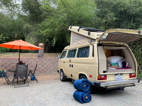 Cozy RV rental setup: A classic Westfalia camper van at a serene campsite with an umbrella, table, and chairs for outdoor living.