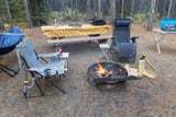 Inviting RV rental campsite featuring a fire pit, hammock, cooking area on a picnic table, and comfy chairs amidst pine trees.