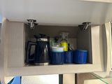 Overhead RV rental kitchen cabinet organized with an electric kettle, blue camping mugs, and cleaning supplies.