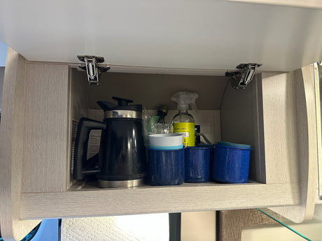 Overhead RV rental kitchen cabinet organized with an electric kettle, blue camping mugs, and cleaning supplies.