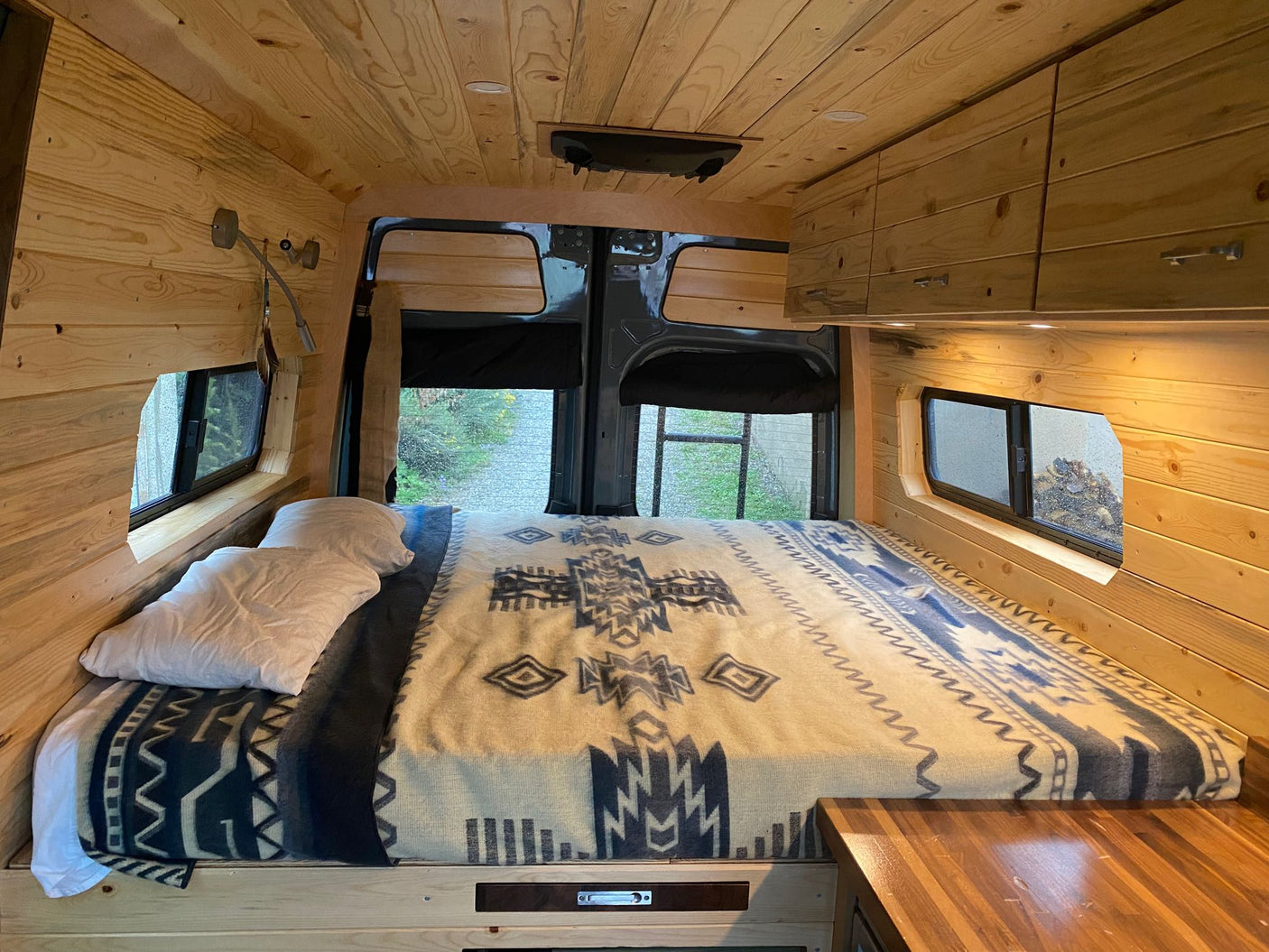 Cozy wood-paneled RV rental camper van interior: Bed with patterned blanket, built-in storage, and rear view.