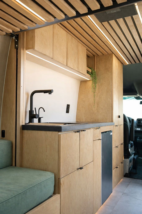 Modern RV rental galley with light wood cabinetry, black sink, fridge, and stylish LED lighting.