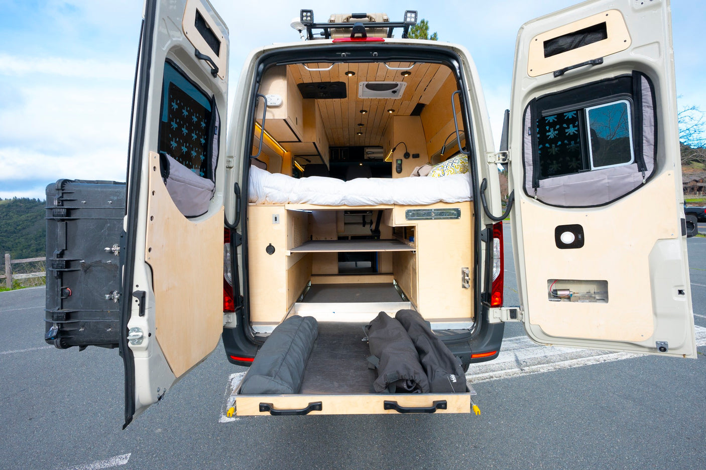 This custom camper van rv rental shows its open rear doors revealing a cozy bed, wooden interior, and pull-out storage for mobile living.