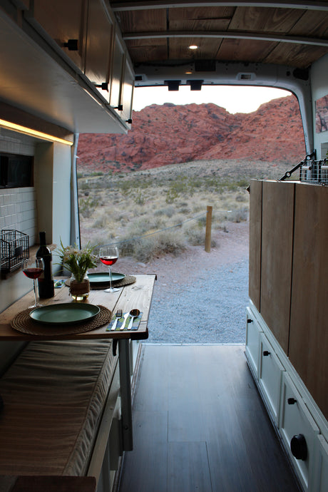 A cozy RV rental camper van interior with a dinner table set for two, overlooking red rock mountains at sunset.