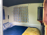 Cozy RV rental interior featuring a comfortable bed with blue bedding, floral curtains, and a compact kitchenette.