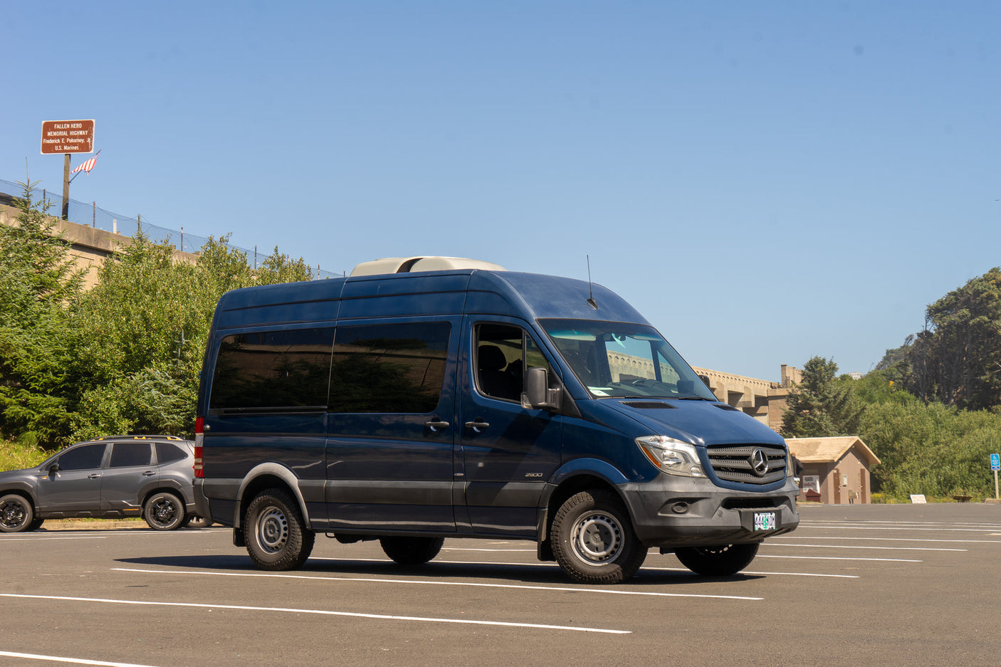 A dark blue Mercedes Sprinter RV rental awaits new adventures in a sunny parking lot, backed by green hills and a large overpass.