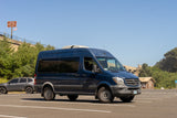 A dark blue Mercedes Sprinter RV rental awaits new adventures in a sunny parking lot, backed by green hills and a large overpass.