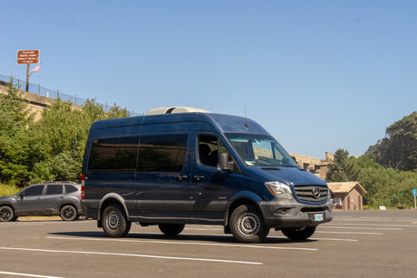 A dark blue Mercedes Sprinter RV rental awaits new adventures in a sunny parking lot, backed by green hills and a large overpass.