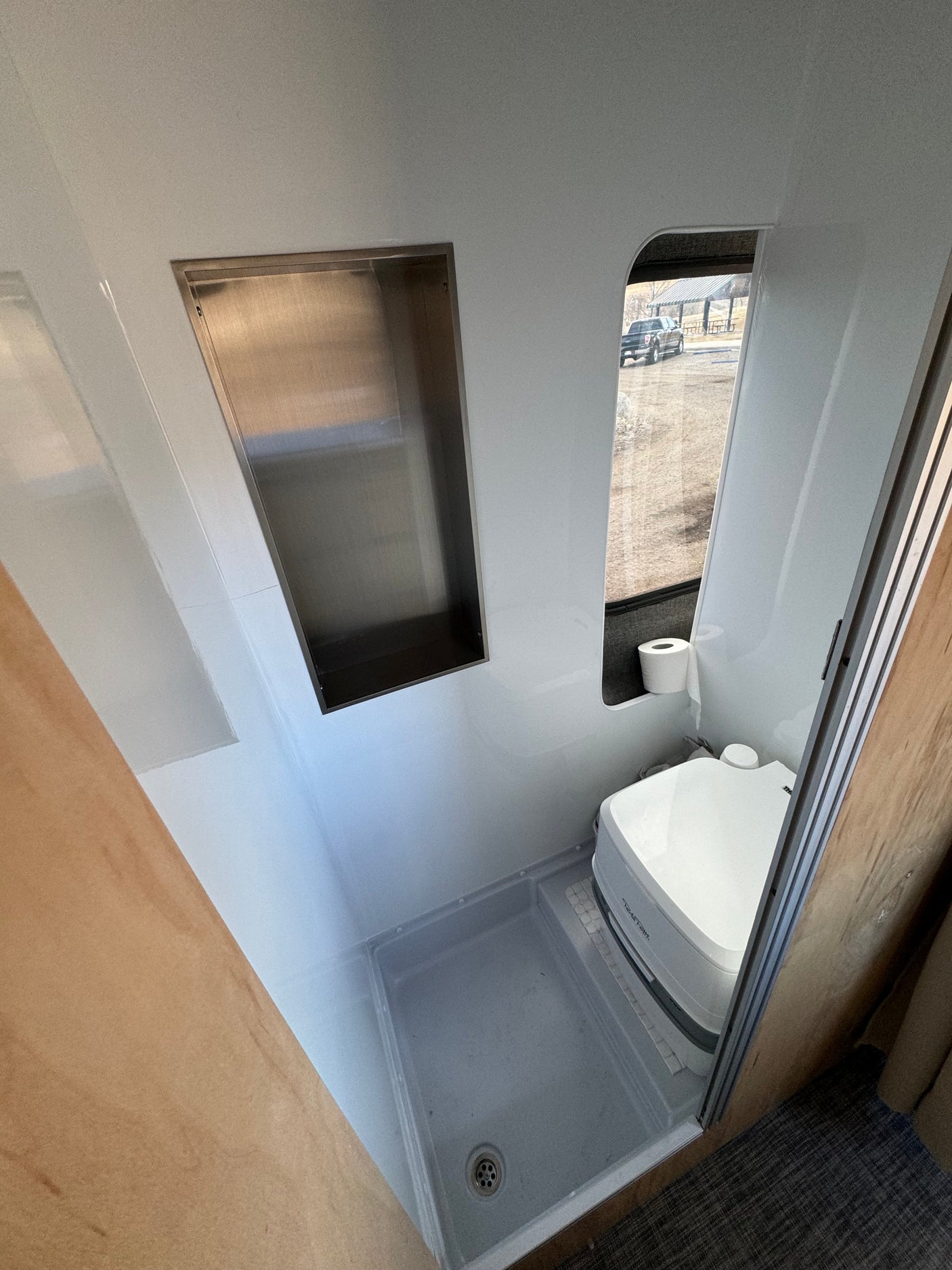 Compact RV rental bathroom with a shower pan, portable toilet, and stainless steel recessed shelf. View out window.