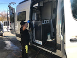 A man cleans an RV rental camper van outdoors, with its side door open revealing a custom interior. Features a rear ladder.