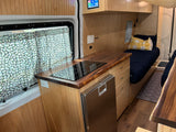 Compact RV rental interior featuring a kitchen with induction cooktop, fridge, and cozy bed area with daisy curtains.