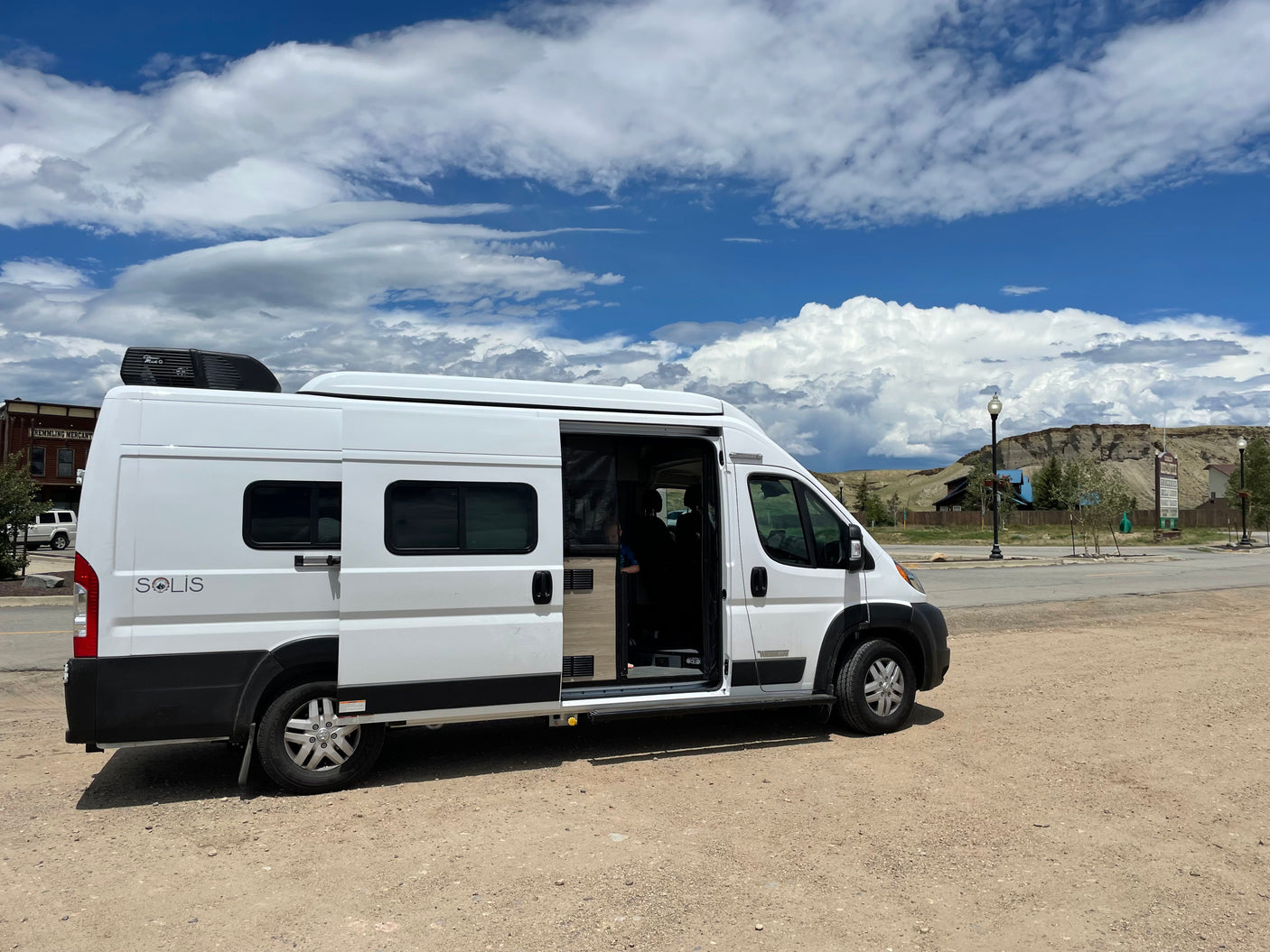 White Winnebago Solis camper van for RV rental, parked on gravel under cloudy sky. Side door open, revealing interior.